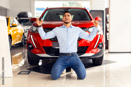 Emotional handsome middle-eastern guy standing on knees, raising hands up, holding key from his brand new red car, copy space. Rich arabic man buying auto at luxury dealership salon, full length photo