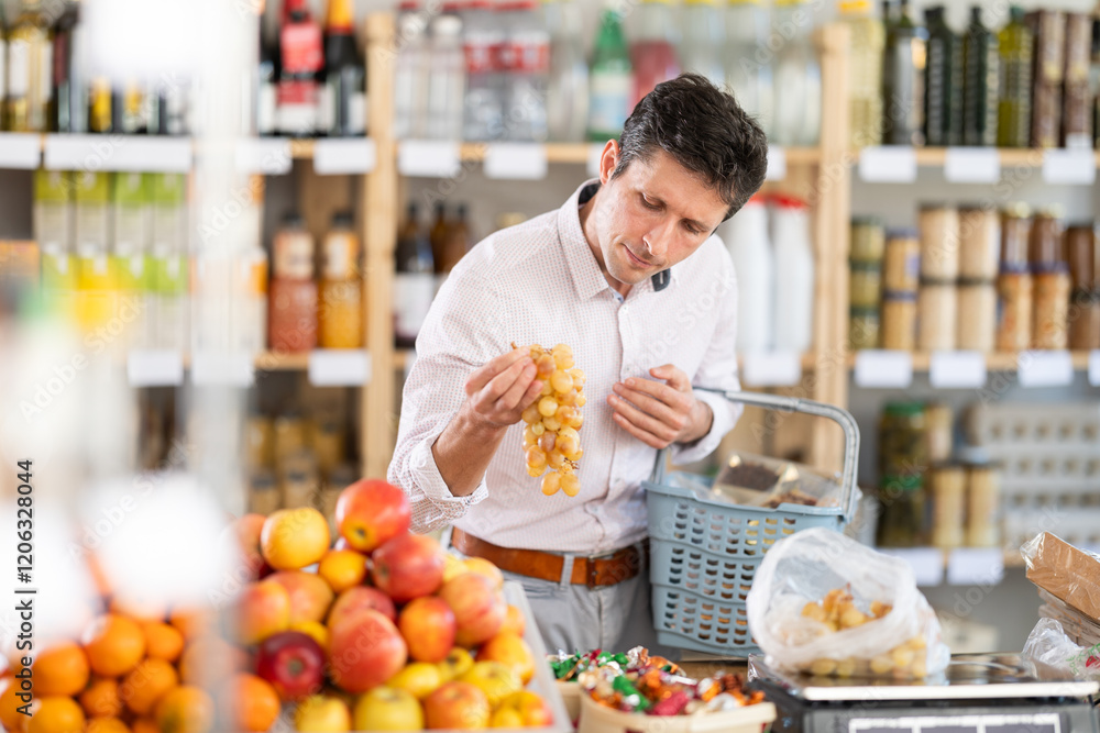 Middle-aged European man standing near fruit stall and choosing ripe Spanish grapes in supermarket Middle-aged European man standing near fruit stall and choosing ripe Spanish grapes in supermarket