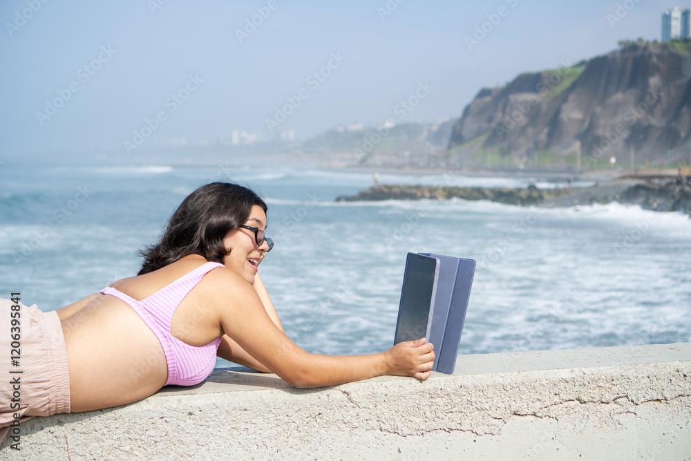 Young woman relaxing on beach wall using tablet, enjoying ocean view Young woman relaxing on beach wall using tablet, enjoying ocean view