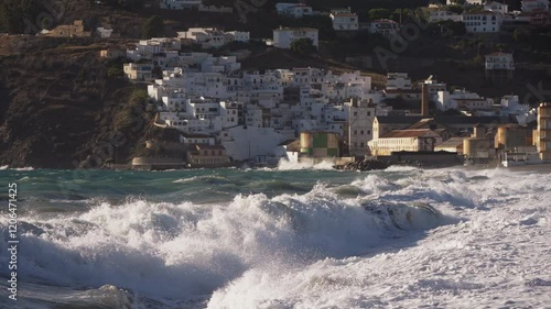 Powerful waves crash against the Mediterranean shoreline, creating a dramatic scene against the backdrop of a charming whitewashed Spanish coastal village. A breathtaking and energetic seascape. photo