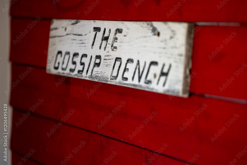 Fototapeta premium A bright red wooden clapboard siding shed wall in a garden with a white wood sign. The words the gossip bench in black letters. The meeting space is for senior retired chatters to sit and gossip.