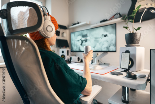 Girl gamer sits comfortably her computer desk, in stylish headphones with microphone, she holds can of drink in her hand and prepares intense video game. Setup enhances focus and gaming experience.