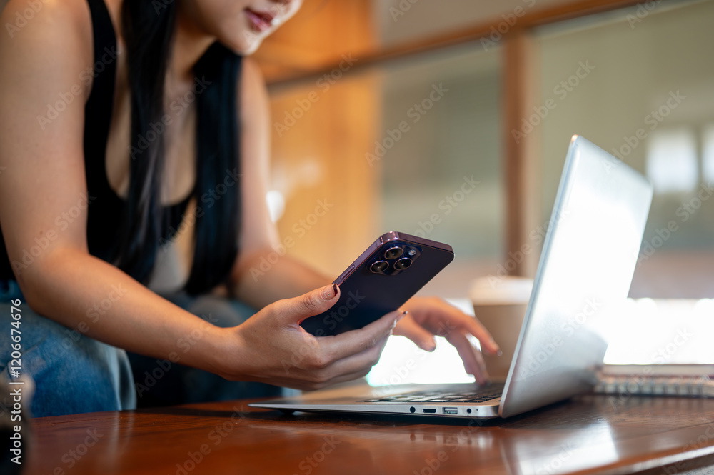 A close-up of an Asian woman using her phone while working remotely on her laptop in a coffee shop. A close-up of an Asian woman using her phone while working remotely on her laptop in a coffee shop.