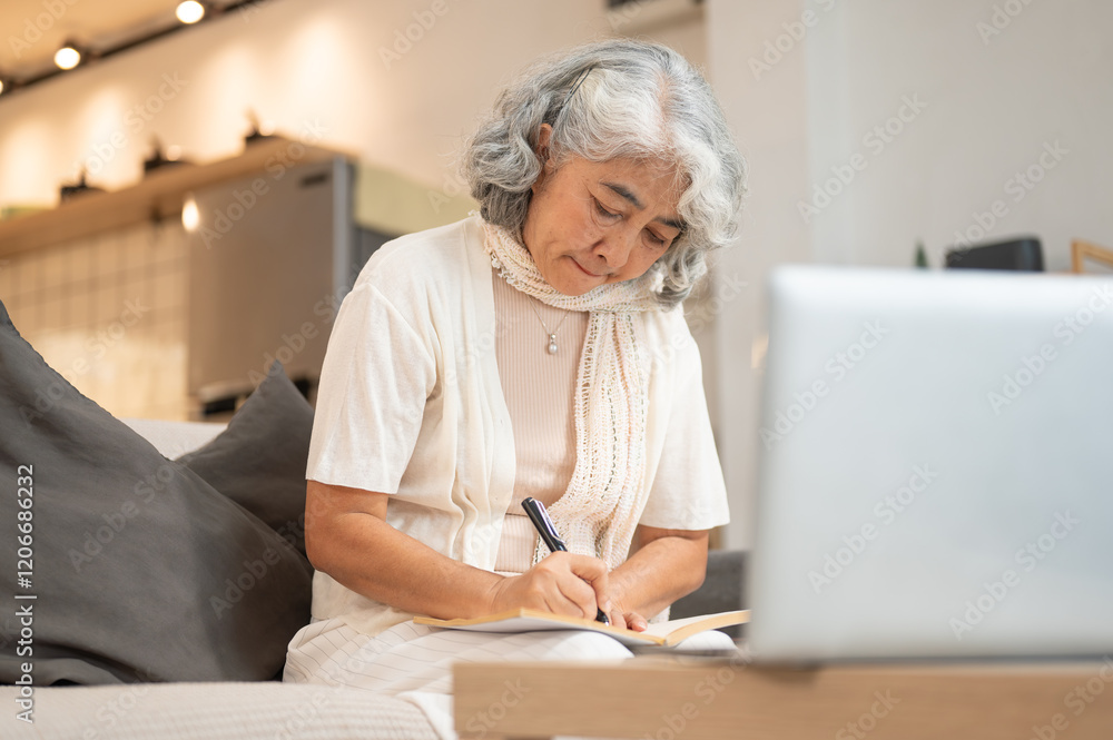 A senior retired woman is focused on studying online at home, writing a lecture in her notebook. A senior retired woman is focused on studying online at home, writing a lecture in her notebook.