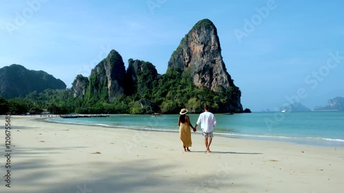 A couple enjoys a leisurely walk on the soft sand of Krabis gorgeous beach, surrounded by towering limestone cliffs and crystal-clear waters. Railay beach photo