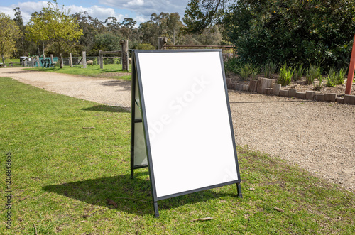 a blank A-frame sandwich board placed on a grassy area in an outdoor park.Empty white background texture with copy space to add custom text or graphics,for promoting events, directions, or information