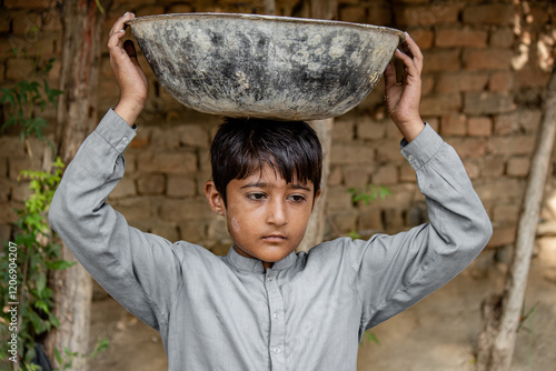 Young boy in traditional gray clothing carrying a large metal bowl on his head, standing outdoors against a rustic brick wall background. photo