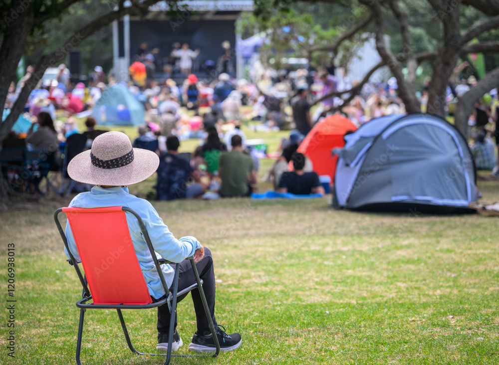 Woman sitting on camping chair, unrecognizable crowds listen to the music concert in the park. Auckland.