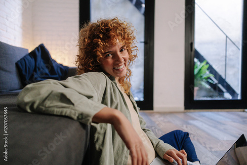 Smiling young woman sitting on floor and holding laptop at home photo