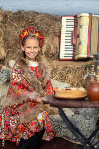 A blonde girl in a national Russian costume on the Maslenitsa holiday. A beautiful russian girl in a national costume made of a fur cape and kokoshnik on the background of a hayloft. 