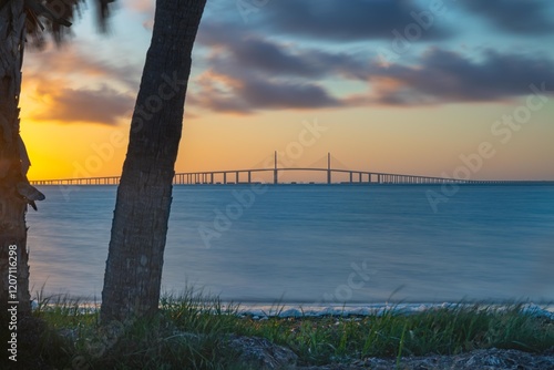 This stunning photograph captures the Sunshine Skyway Bridge from Fort DeSoto Park at sunrise, where the first light of day casts a warm glow across the tranquil waters. The soft light the golden hr