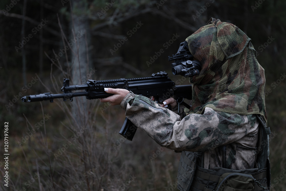 Fototapeta premium A young girl special forces soldier in a gas mask, camouflage and a tactical helmet with a night vision monocular in the forest at dusk.