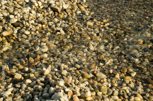 Pebbles and stones beneath the shallow surface of crystal-clear water. The sunlit scene creates shimmering reflections highlighting the textures and colors of the stones.