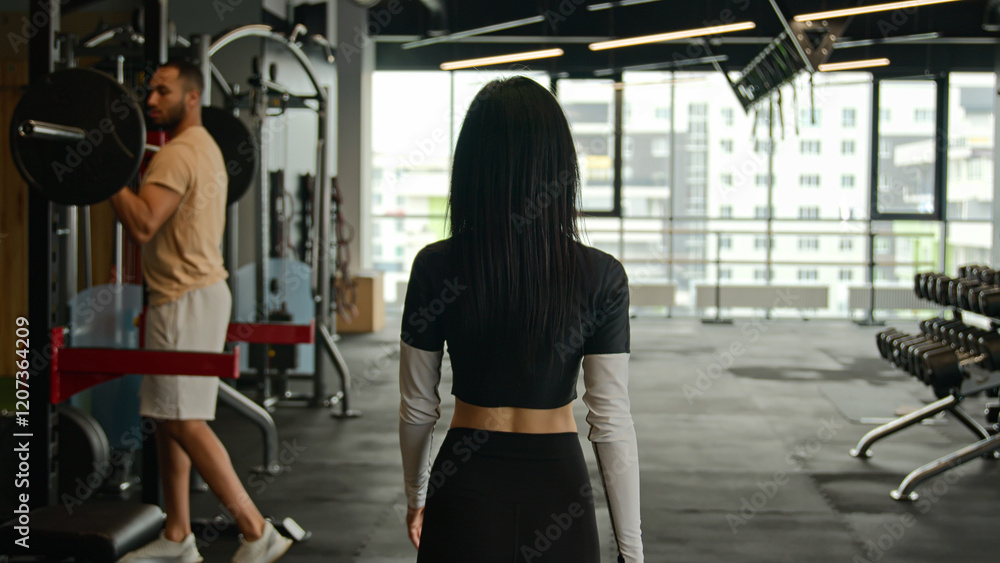 Back view from behind unrecognizable woman fitness trainer sportswoman girl going walking in sports club in modern gym african american man sportsman training doing barbell exercise sport equipment Back view from behind unrecognizable woman fitness trainer sportswoman girl going walking in sports club in modern gym african american man sportsman training doing barbell exercise sport equipment