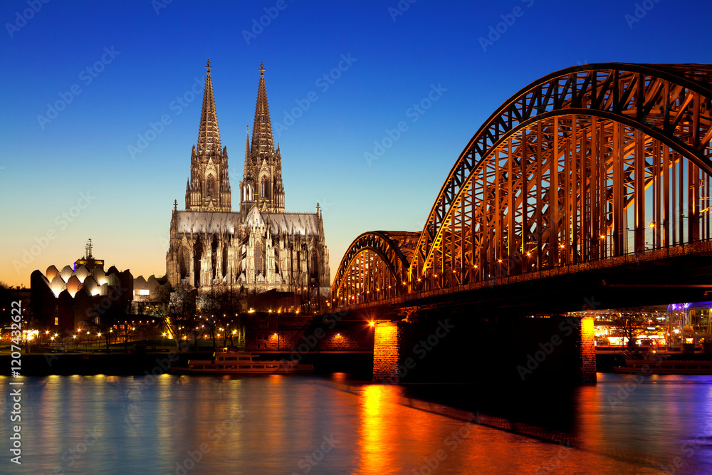Fototapeta premium Kölner Dom und Hohenzollernbrücke bei Nacht – Beleuchtete Skyline von Köln mit Spiegelungen im Rhein