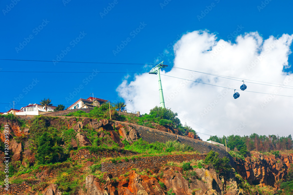 Cable cars glide above a lush, rocky hillside under a bright blue sky with fluffy clouds. A scenic view featuring rugged terrain, vibrant greenery, and charming hilltop houses