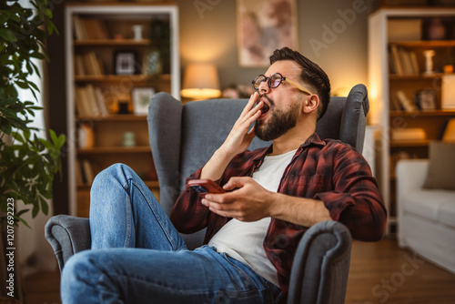 sleepy adult man sit in the armchair yawning and use mobile phone photo