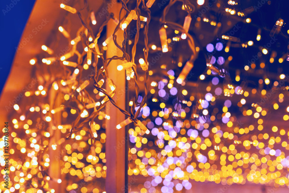 String lights hanging from a structure, with a background filled with bokeh effects of various lights. Warm yellow and purple hues create a festive and visually appealing atmosphere