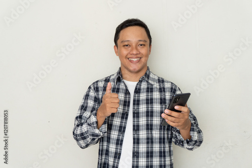 Young Asian man smiling happy and showing thumbs up gesture while holding mobile phone. photo