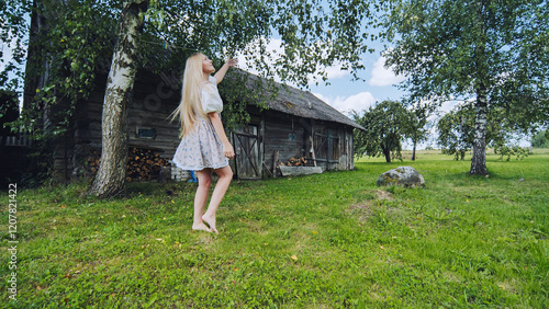 Barefoot woman stretching toward wooden barn branch, long blond hair flowing under summer sunlight photo