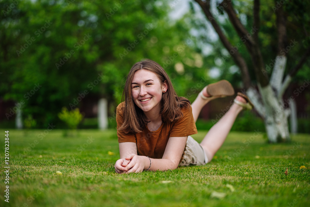 Cheerful teen relaxing on green lawn, grinning widely amid blooming garden during sunny summer afternoon Cheerful teen relaxing on green lawn, grinning widely amid blooming garden during sunny summer afternoon