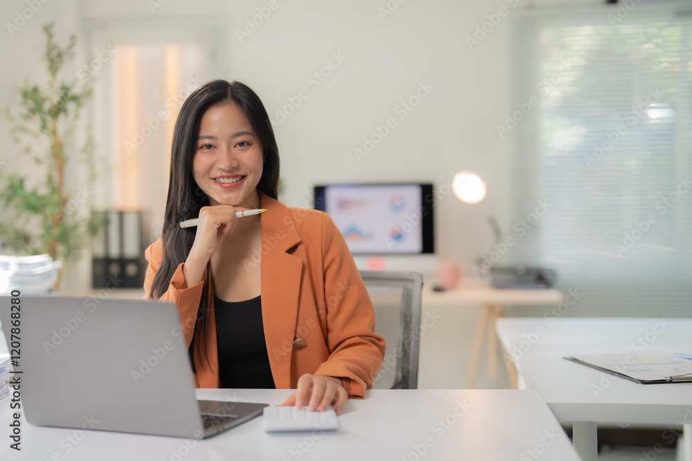 Young happy Asian businesswoman in orange suit smiling while working using calculator and laptop in modern office, with computer showing charts in background Young happy Asian businesswoman in orange suit smiling while working using calculator and laptop in modern office, with computer showing charts in background