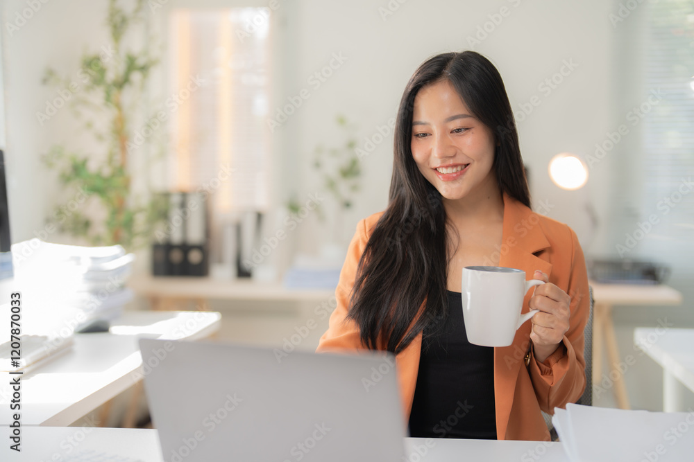 Young Asian manager enjoying a hot cup of coffee while working on her laptop, creating a cheerful and productive atmosphere in a modern office setting Young Asian manager enjoying a hot cup of coffee while working on her laptop, creating a cheerful and productive atmosphere in a modern office setting