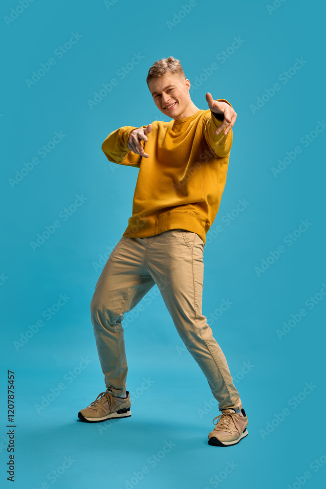 Man pointing forward with playful smile, leaning slightly, engaging expressively with energetic gestures against blue studio background. Concept of human emotions, beauty, self-expression. Man pointing forward with playful smile, leaning slightly, engaging expressively with energetic gestures against blue studio background. Concept of human emotions, beauty, self-expression.