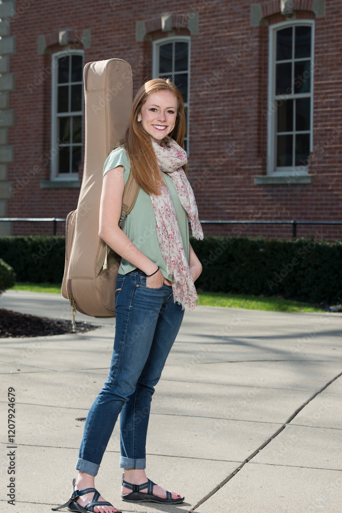 Fototapeta premium Beautiful young redhead woman on campus carrying a guitar case - smiling