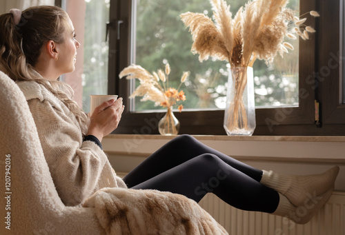 Junge Frau schaut aus dem Fenster, trinkt Tee und legt Füße zum Aufwärmen auf die Heizung