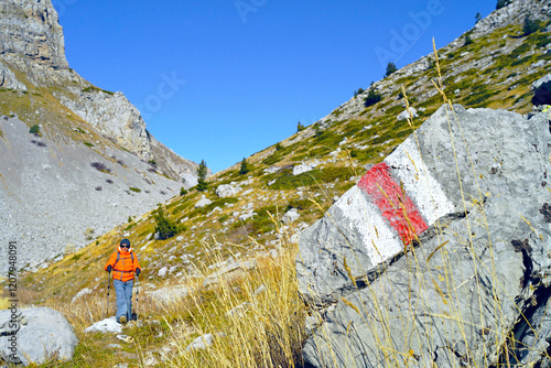 Hiking routes in the mountains of Albania: a close-up photograph of a rock with red and white tourist markings and a hiker walking along the trail in the background. Male tourist in Buni i Jezerces photo