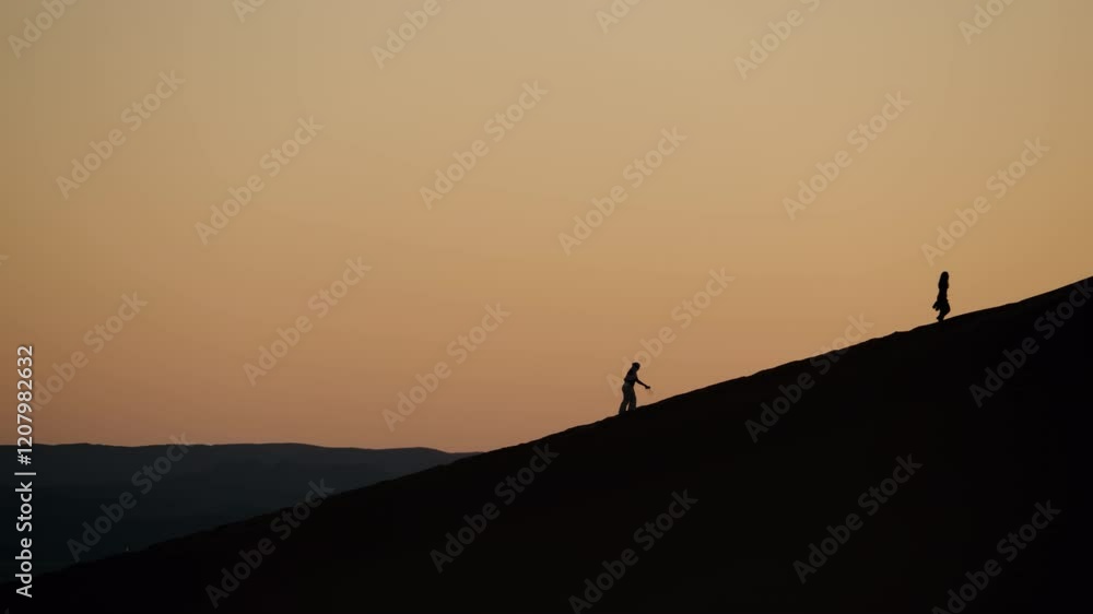 Silhouetted figures climb a sand dune at sunset in the Moroccan desert. The warm colors of the sky create a tranquil atmosphere. This serene scene evokes feelings of adventure, travel, exploration. Silhouetted figures climb a sand dune at sunset in the Moroccan desert. The warm colors of the sky create a tranquil atmosphere. This serene scene evokes feelings of adventure, travel, exploration.