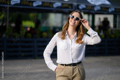 Stylish woman standing on city street in fashionable sunglasses and snow-white shirt, smiling happily and enjoying sunny day. Travel, dream concept