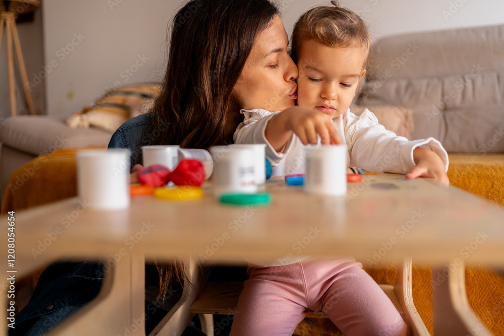 Mom kissing her little daughter playing with modeling clay at home, having fun and learning Mom kissing her little daughter playing with modeling clay at home, having fun and learning