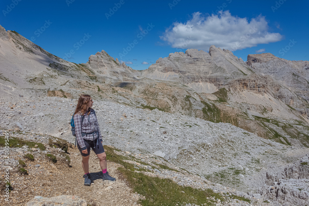 Fototapeta premium Young woman with long hair looking at the awesome summer valley panorama in the Dolomites, Mount Latemar, Trentino, Italy. Traveling photography and outdoor sport activity concept