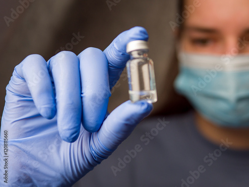 Doctor wearing surgical mask and blue latex gloves holding medical vial containing clear liquid, carefully preparing for medical procedure, emphasizing safety with medical vial in focus. 