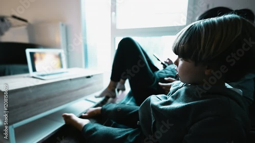 Small boy hypnotized by cartoon seated at home sofa. Child watching entertainment media on couch while mother using cellphone. Technology addiction concept