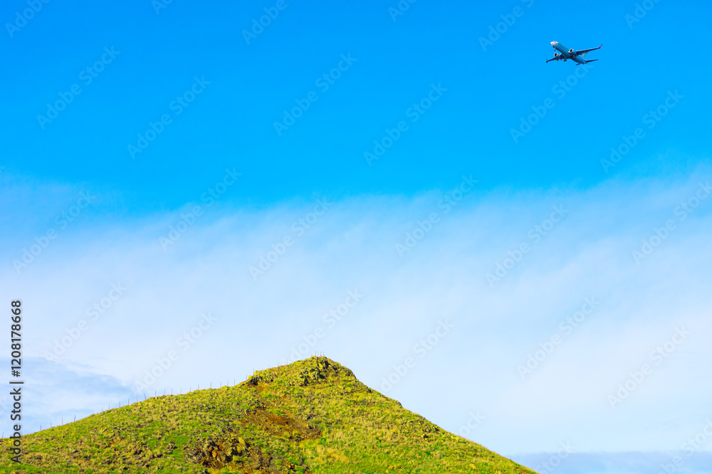 Green hill with a rocky top under a bright blue sky, with a commercial airplane flying above it. Contrast between natural landscape and the aircraft creates an interesting visual juxtaposition