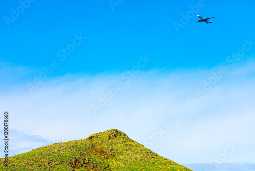 Obraz na płótnie Green hill with a rocky top under a bright blue sky, with a commercial airplane flying above it. Contrast between natural landscape and the aircraft creates an interesting visual juxtaposition w salonie