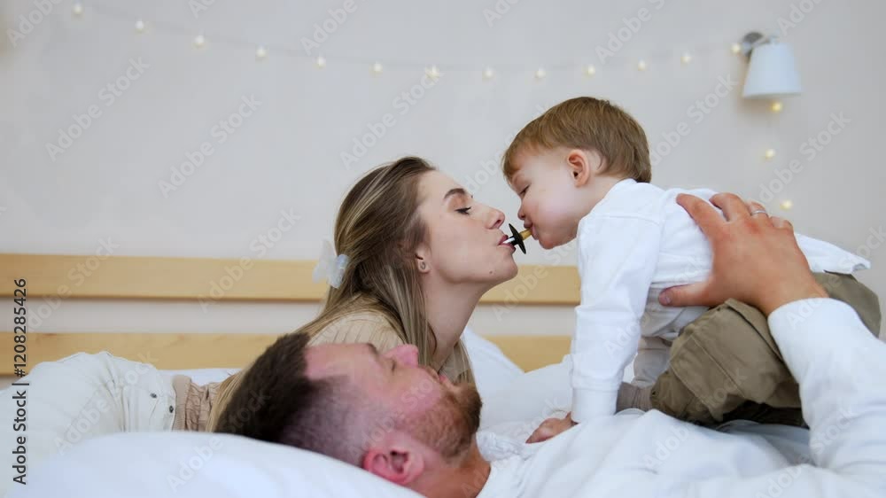 Cute baby boy sitting on his dad's belly. Mom is holding a pacifier in her mouth and toddler kid tries to get it. Family resting in the bedroom together.