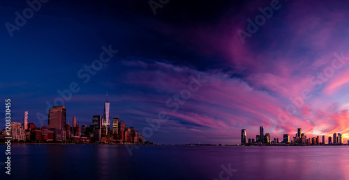 Panoramic view of Lower Manhattan and Jersey City during a vibrant sunset. The sky is painted with dramatic hues of purple, pink, and orange, reflecting softly on the calm waters of the Hudson River.