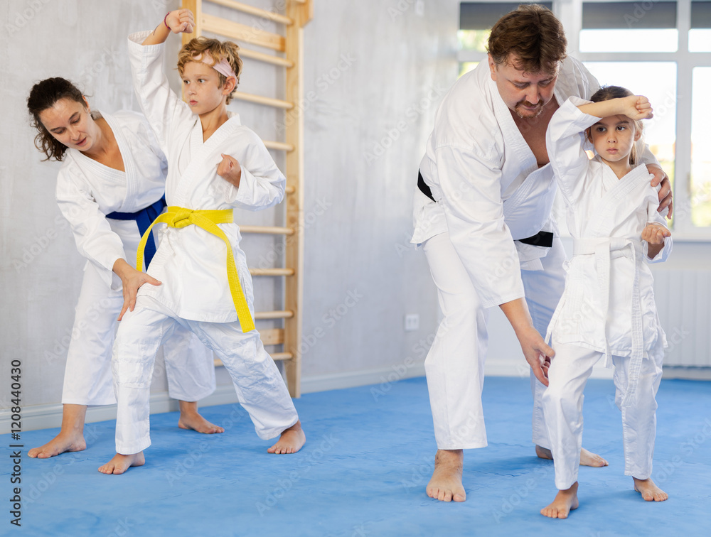 Fototapeta premium Concentrated preteen boy and girl in white kimonos diligently performing kata exercises to hone martial arts skills under guidance of experienced parents. Concept of family joint sports activity