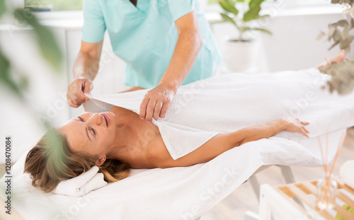 Pleased middle-aged woman lying in physiotherapy room while elderly masseuse wrapping her with white coverlid photo