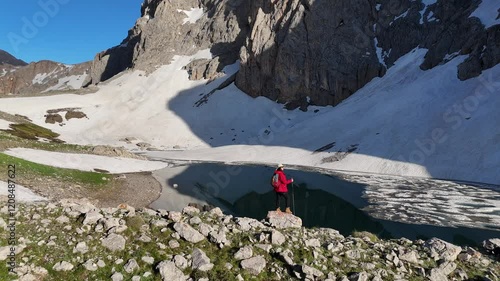 An adventurous hiker, a tourist woman on a mountain hike, standing on a rock near a serene alpine lake surrounded by snow-covered landscapes, reflecting the breathtaking beauty of untouched nature. photo