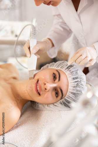 Interested young female patient undergoing facial skin cleansing with ultrasonic shovel to enhance efficacy of skincare products and improving skin texture. Modern hardware cosmetology photo