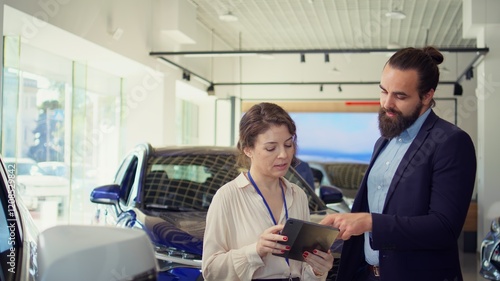 Customer in showroom looking for new vehicle to purchase, being shown option by saleswoman on tablet. Agent using device to help man evaluate automobiles in dealership before buying, camera B photo