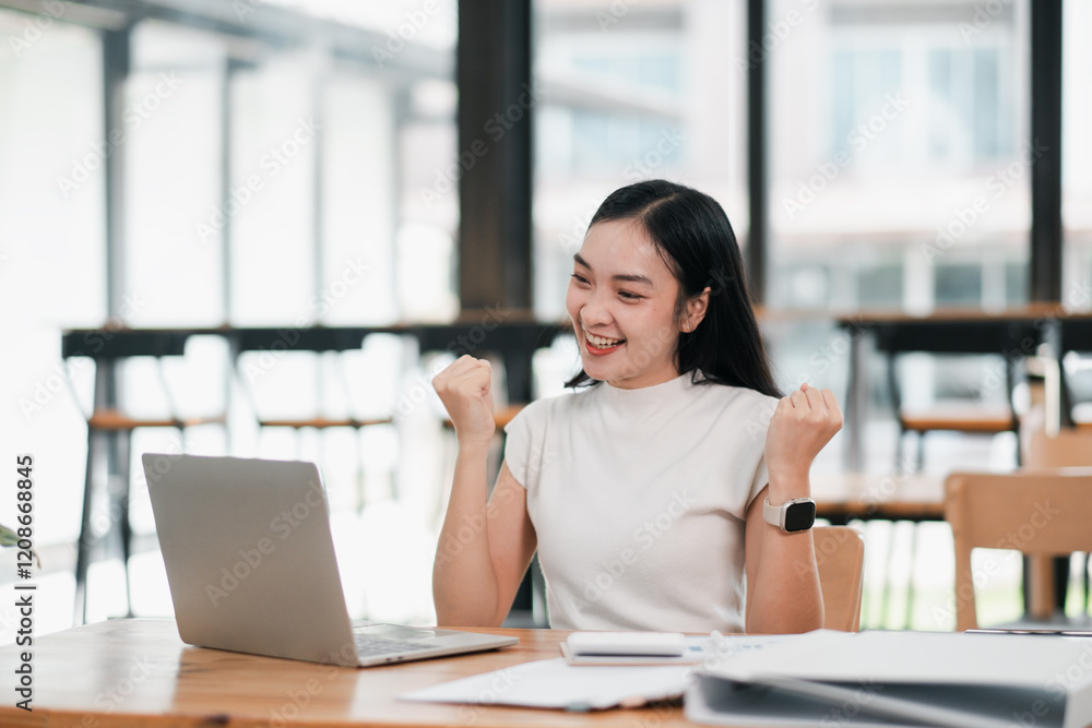 A joyful young woman celebrates success while working on her laptop in a bright, modern office setting. A joyful young woman celebrates success while working on her laptop in a bright, modern office setting.