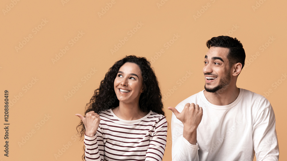 Joyful Arab Man And Woman Looking Aside And Pointing At Copy Space With Thumbs Up, Interested Middle Eastern Couple Sitting On Floor Over Yellow Studio Background And Showing Free Place For Ad Joyful Arab Man And Woman Looking Aside And Pointing At Copy Space With Thumbs Up, Interested Middle Eastern Couple Sitting On Floor Over Yellow Studio Background And Showing Free Place For Ad