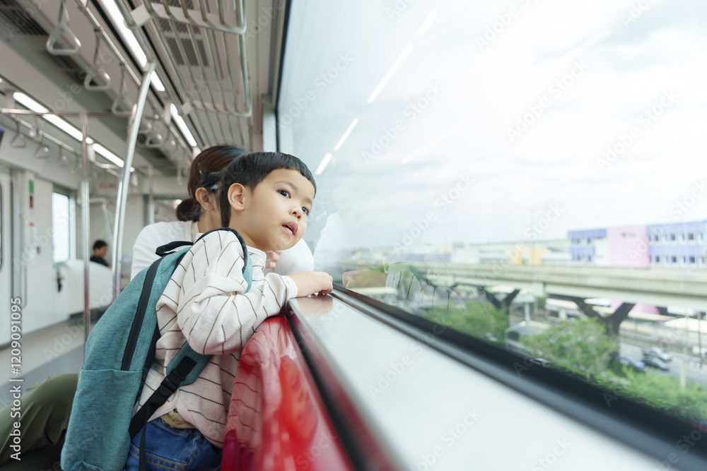 Fototapeta premium Asian boy with a backpack looking out the window of an elevated train while going to the school, accompanied by his mother, enjoying the city view during travel.