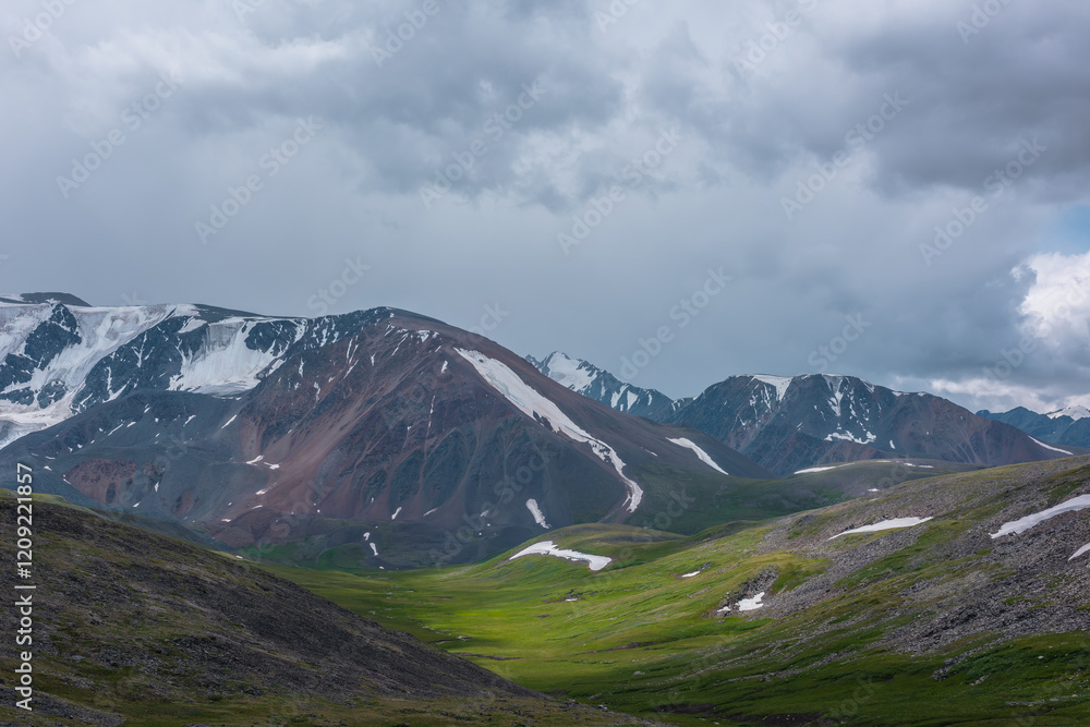 Dramatic overcast view to beautiful green alpine valley among stony and grassy hills, big snowy range and part of mountain with snow-capped sharp pinnacle far away in center under rain gray cloudy sky Dramatic overcast view to beautiful green alpine valley among stony and grassy hills, big snowy range and part of mountain with snow-capped sharp pinnacle far away in center under rain gray cloudy sky